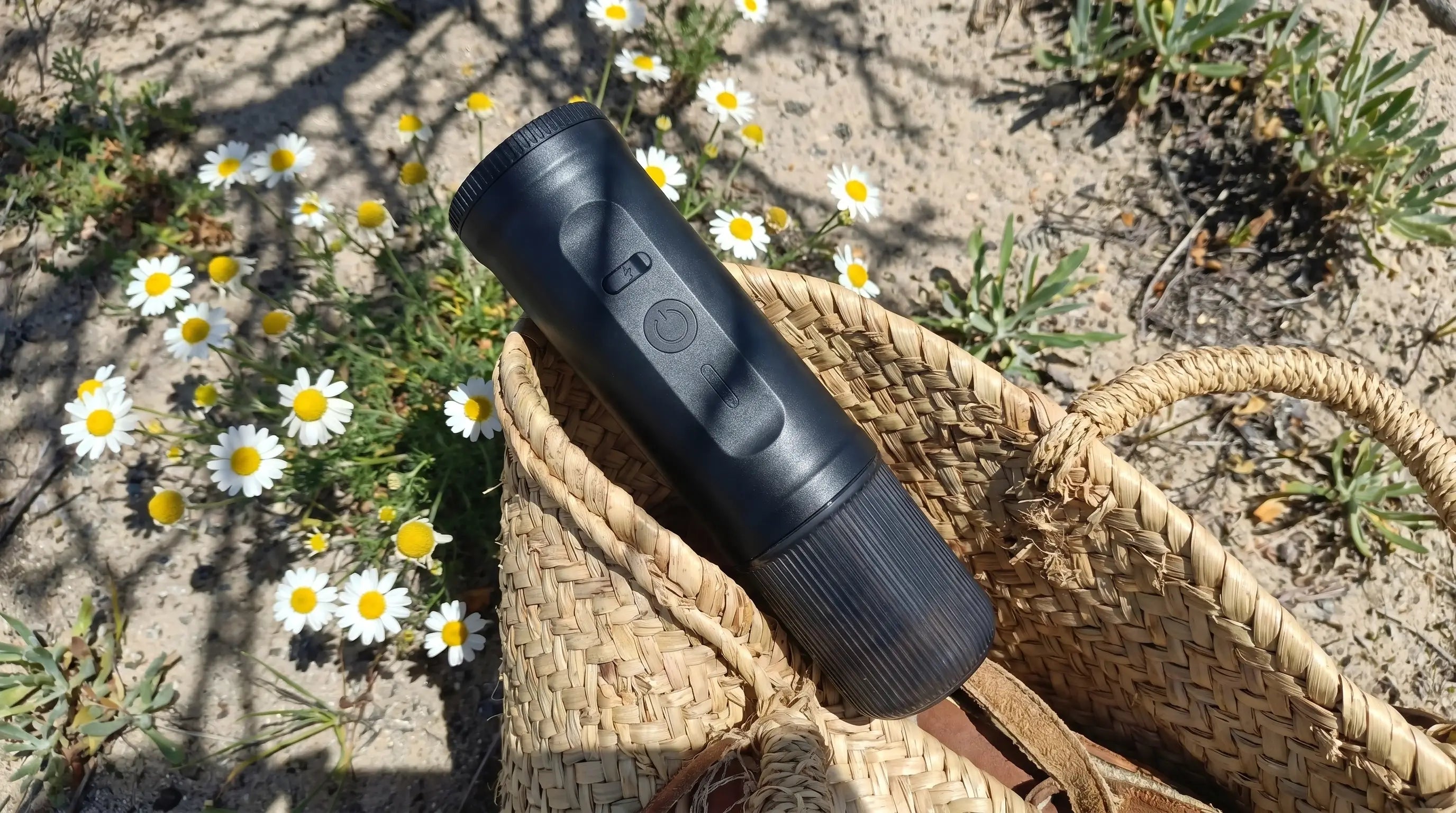 Yominobo portable espresso machine resting in a wicker basket among daisy flowers outdoors — compact and lightweight design for outdoor brewing