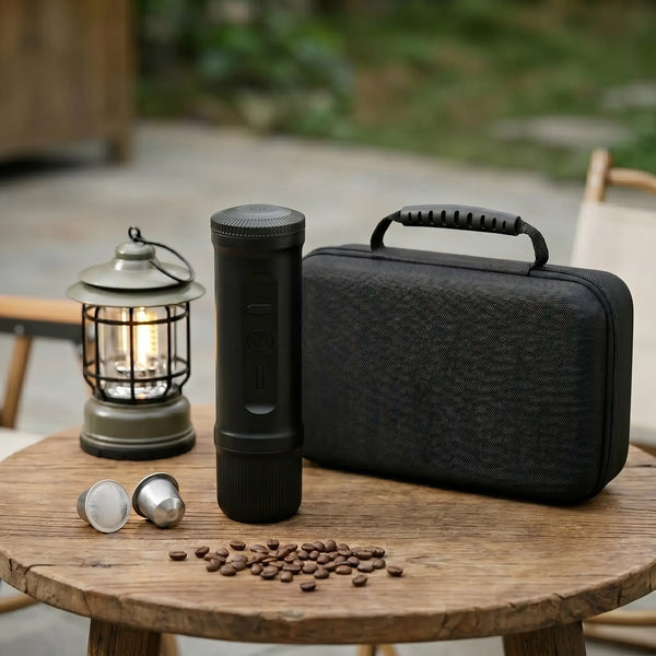 YomiNobo matte black cylindrical portable espresso machine with textured anti-slip ribs, standing on a wooden camp table next to two capsules, loose beans, a black hardshell travel case, and a vintage-style camp lantern in an outdoor leisure setting.