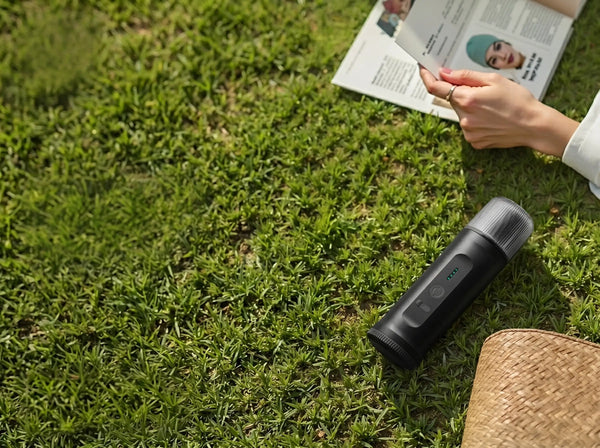High-angle shot of a YomiNobo matte black portable espresso machine resting on a lush green lawn, with a person reading a magazine and a straw bag in a relaxing outdoor leisure setting.